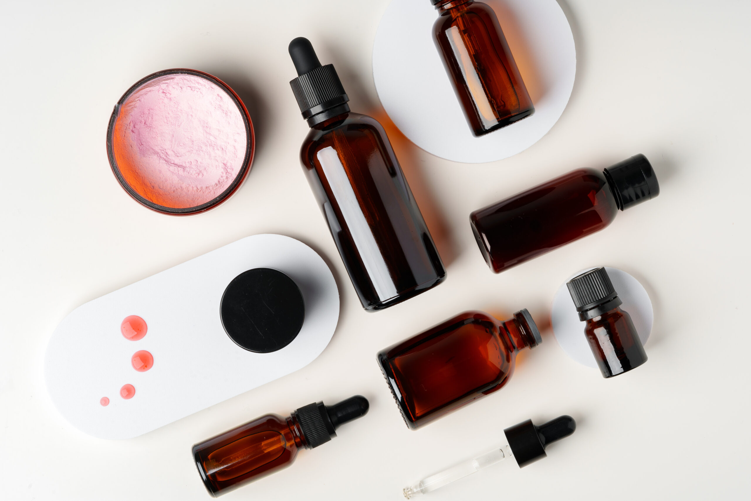 A collection of amber glass bottles and jars containing skincare products are arranged on a white background. The bottles are various shapes and sizes, some with droppers, some with lids, and some with a pump. There are also a few drops of red liquid on the surface.